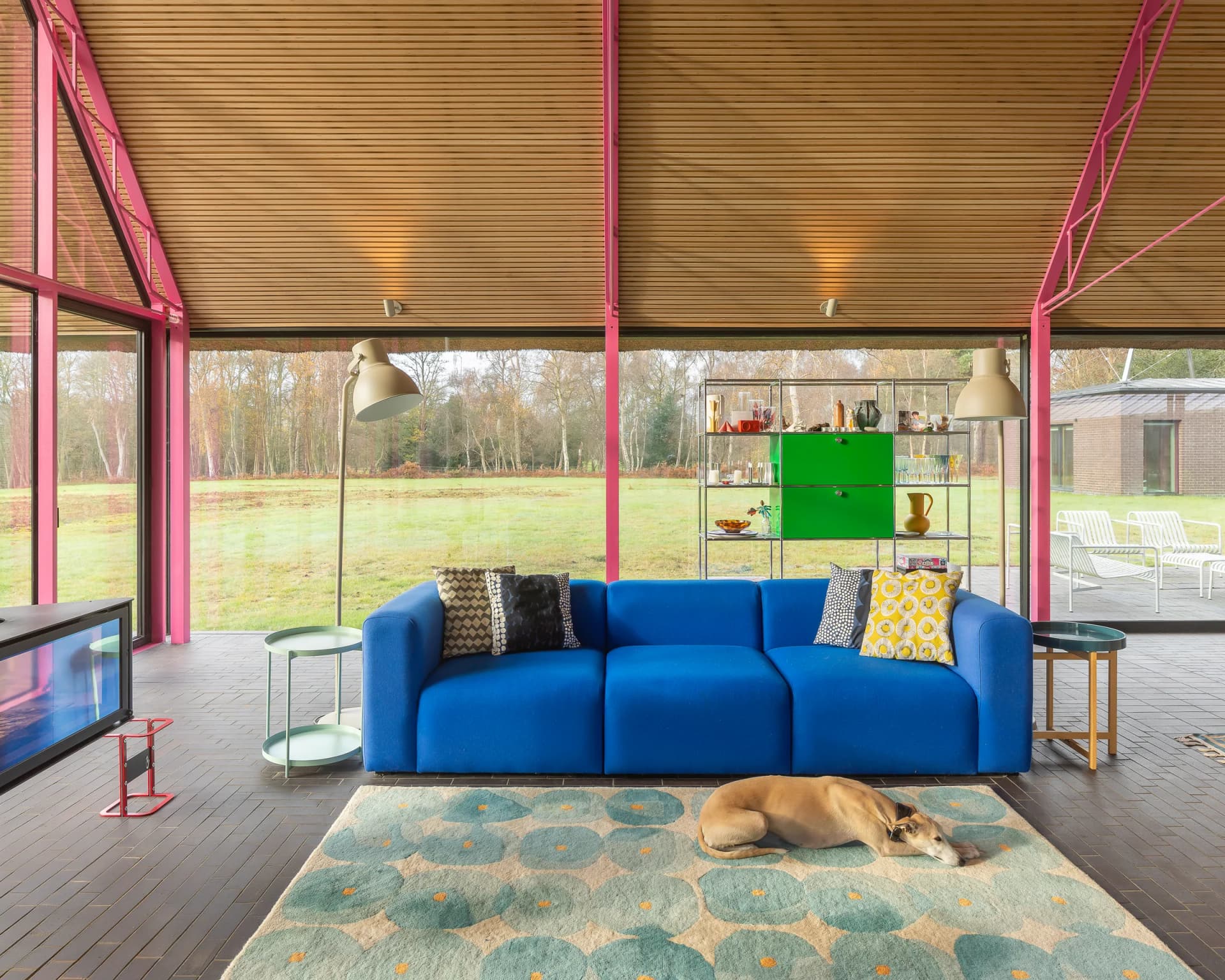 Inside Housestead: a timber-lined living space framed by exposed pink steel structure and full-height glazing wrapping the corner to the Suffolk landscape.