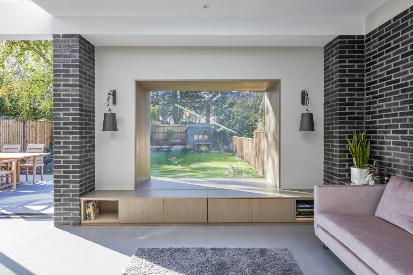 Interior view through a Maxlight Oriel Bay Window: an oak-lined projecting bay with frameless glass-to-glass corners framing a garden, set between brick walls above a low timber bench.