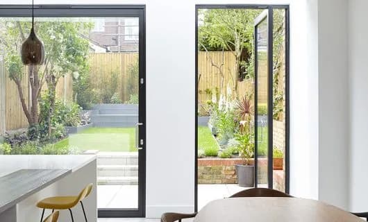 Framed pivot door open from a dining space towards a garden