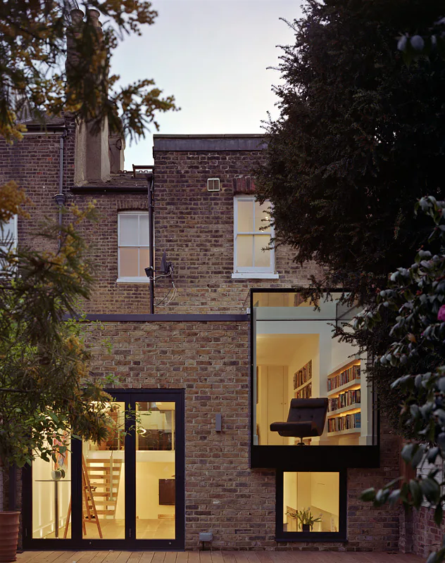 Projecting glass reading room glowing on the rear of a brick house at dusk