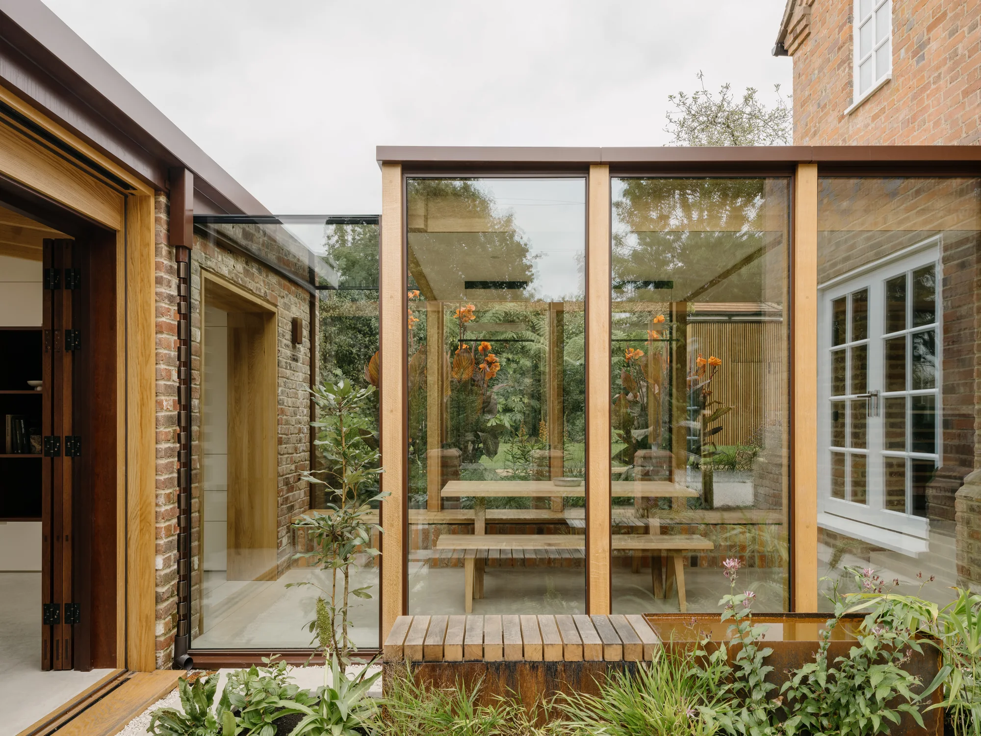 Full-height glazing with warm metal frames enclosing a courtyard beside a brick house
